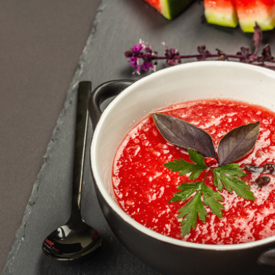 Cuenco de gazpacho de sandía frío decorado con hierbas frescas, junto a trozos de sandía sobre una superficie oscura.