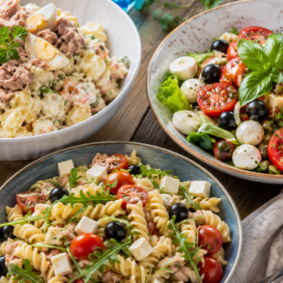Tres ensaladas de verano servidas sobre una mesa de madera al aire libre: una ensaladilla rusa tradicional, una ensalada mediterránea con mozzarella y tomates cherry y una ensalada de pasta, ideales para los días más calurosos del año.